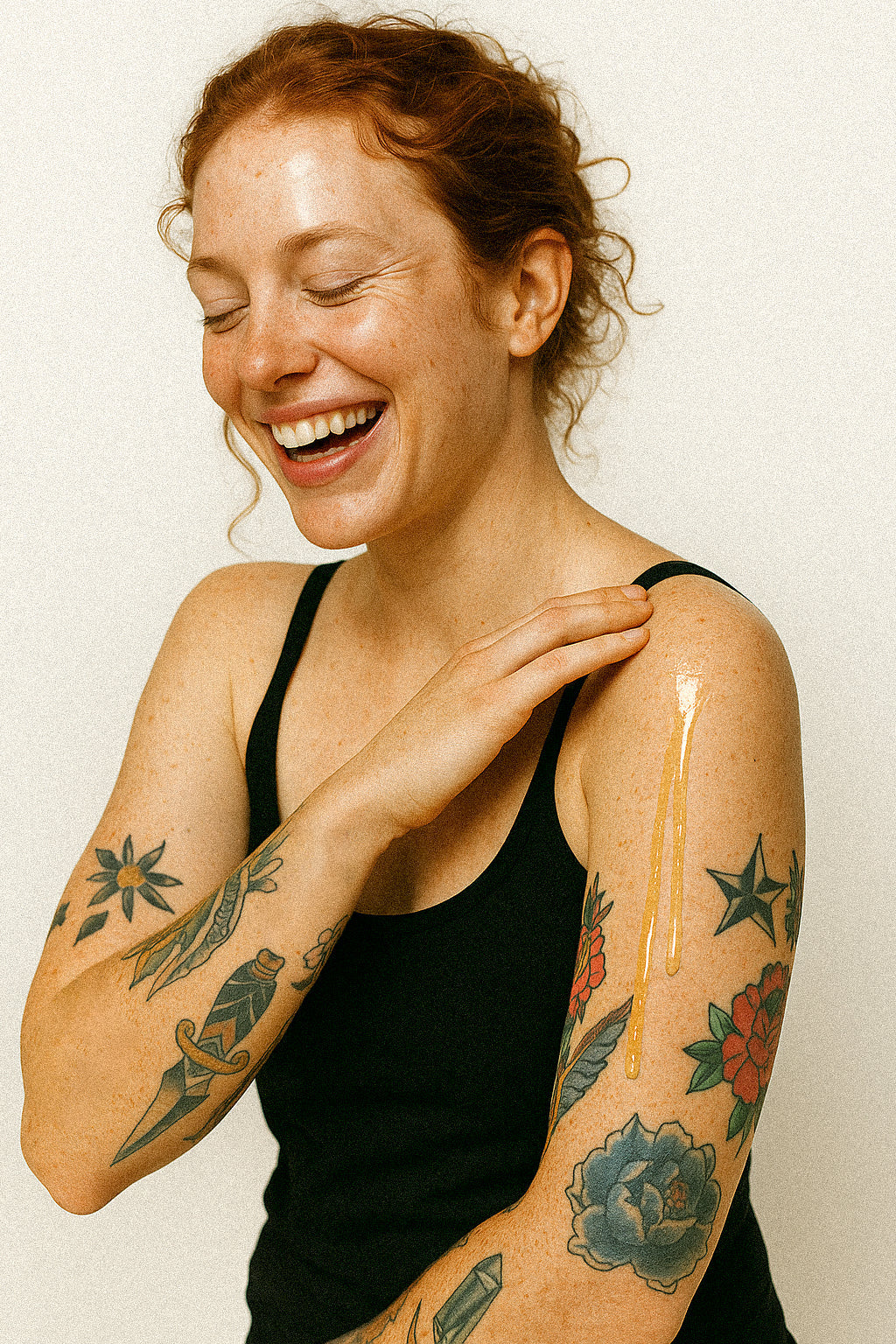 Woman with tattoos on her arms, smiling against a plain background with non toxic body oil running down arm