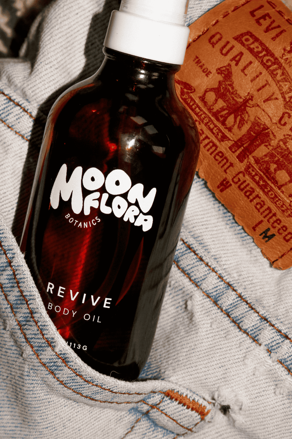 Bottle of moonflora botanics Revive organic Body Oil on denim fabric with a Levi's tag.
